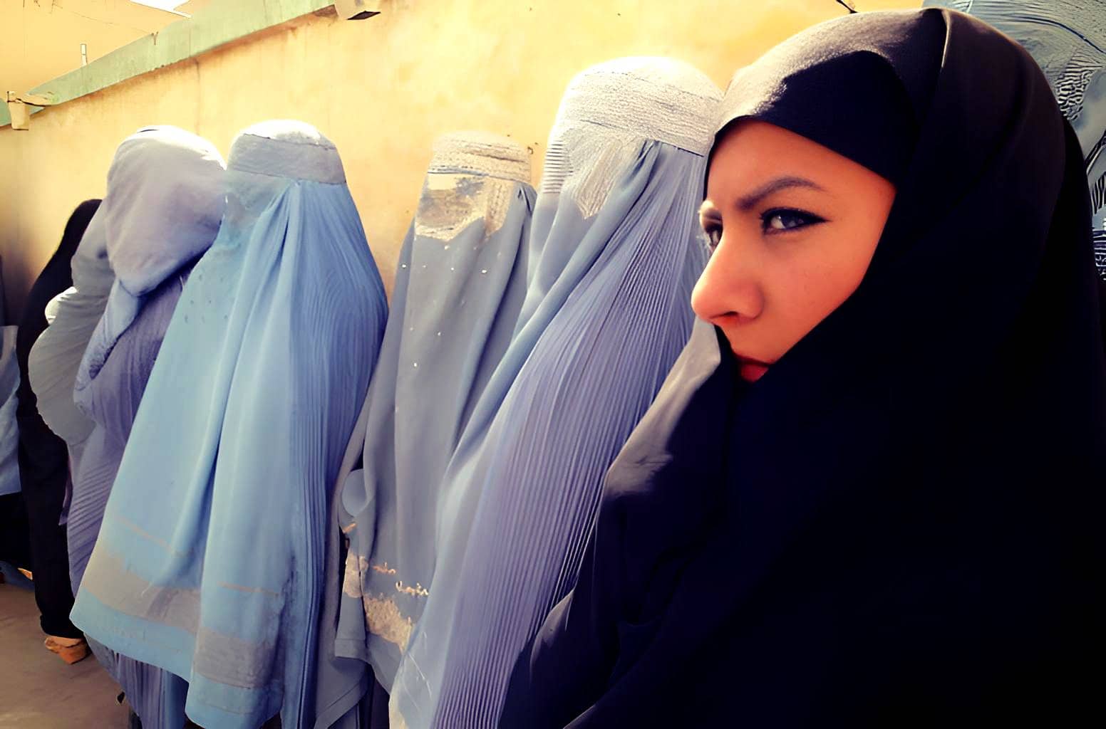 Brightly dressed women wearing traditional blue and white garments, standing in a row, with one woman prominently visible wearing a black headscarf, highlighting cultural attire and community gathering.