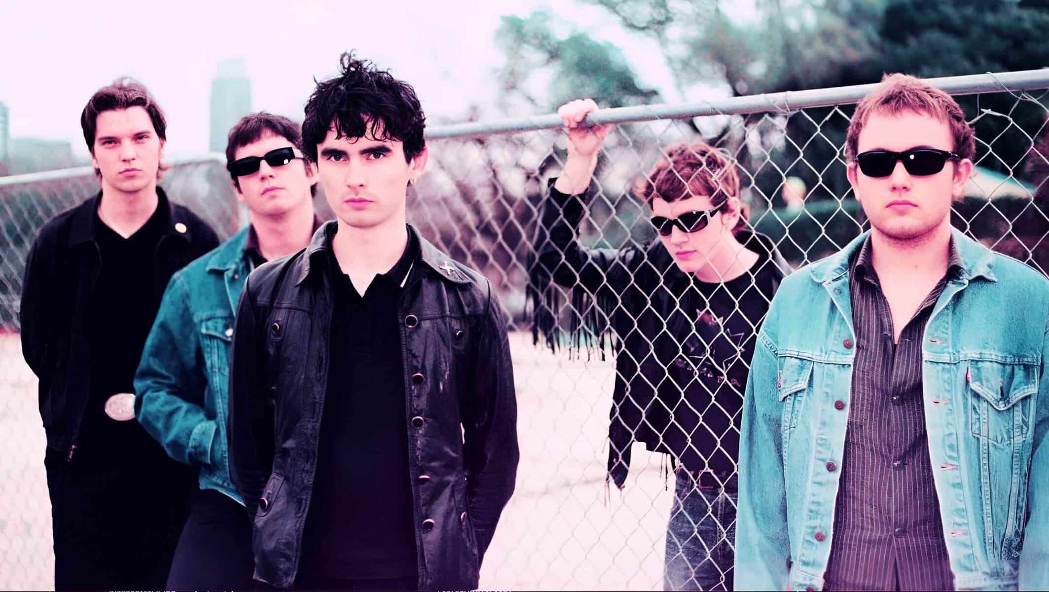 Vintage rock band members standing near a chain-link fence during a music photoshoot.