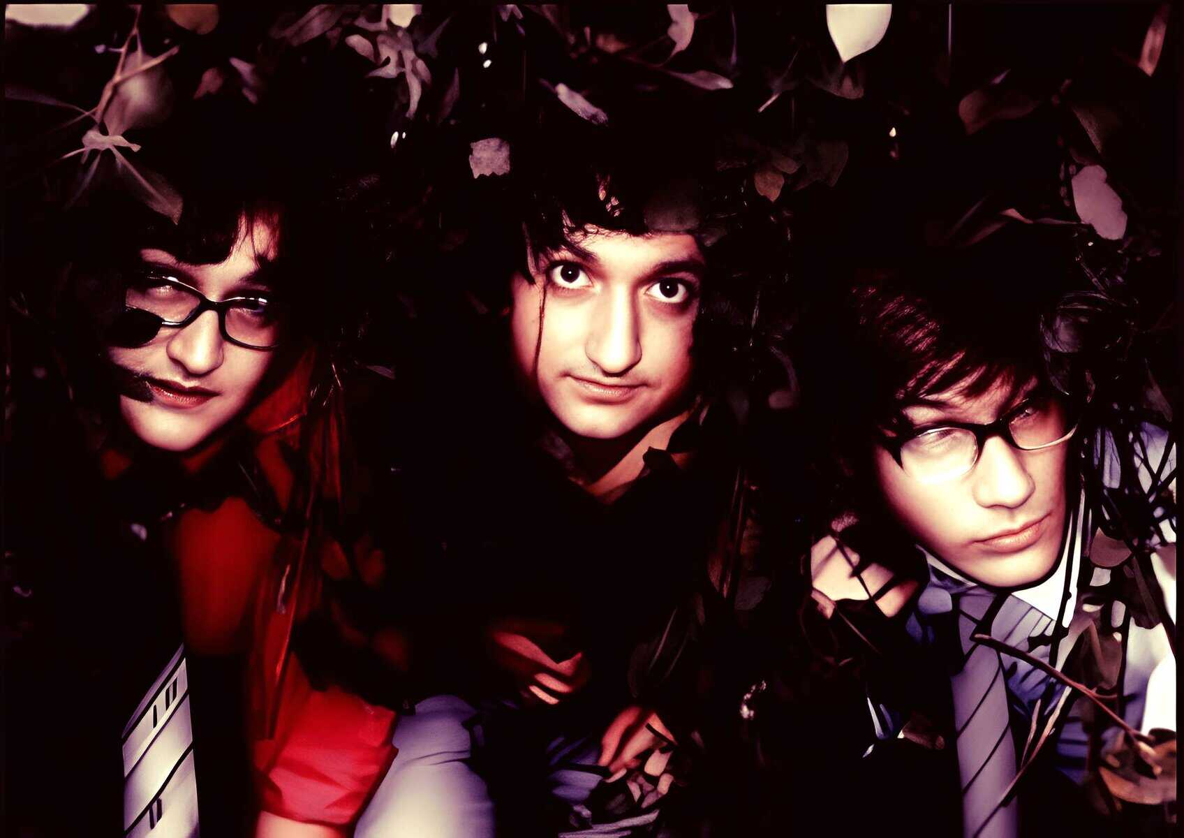 Hidden amid dark foliage, three young adults with distinctive styles look directly at the camera, showcasing contemporary music themes and alternative fashion often featured in modern sound archive collections.