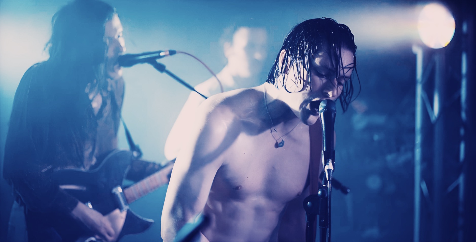 Vibrant live performance of a shirtless male singer and guitarist passionately singing into microphone at a music concert, with bandmates and stage lights in the background, capturing the essence of live music and rock performances.