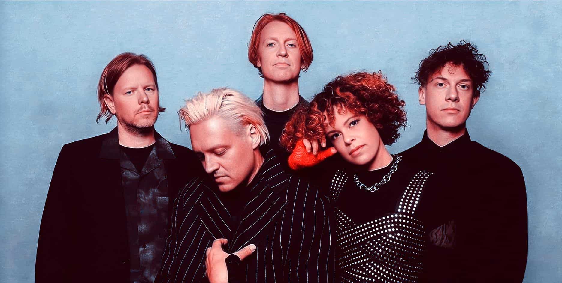 Vintage rock band photo with five members dressed in stylish black outfits, showcasing 80s music culture and iconic band style, against a textured light blue background.