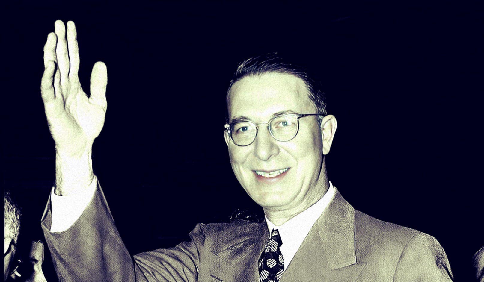 A man in a suit and tie waving, with a smile, wearing glasses, representing a historical news or music archive context for past daily sound recordings and media preservation.