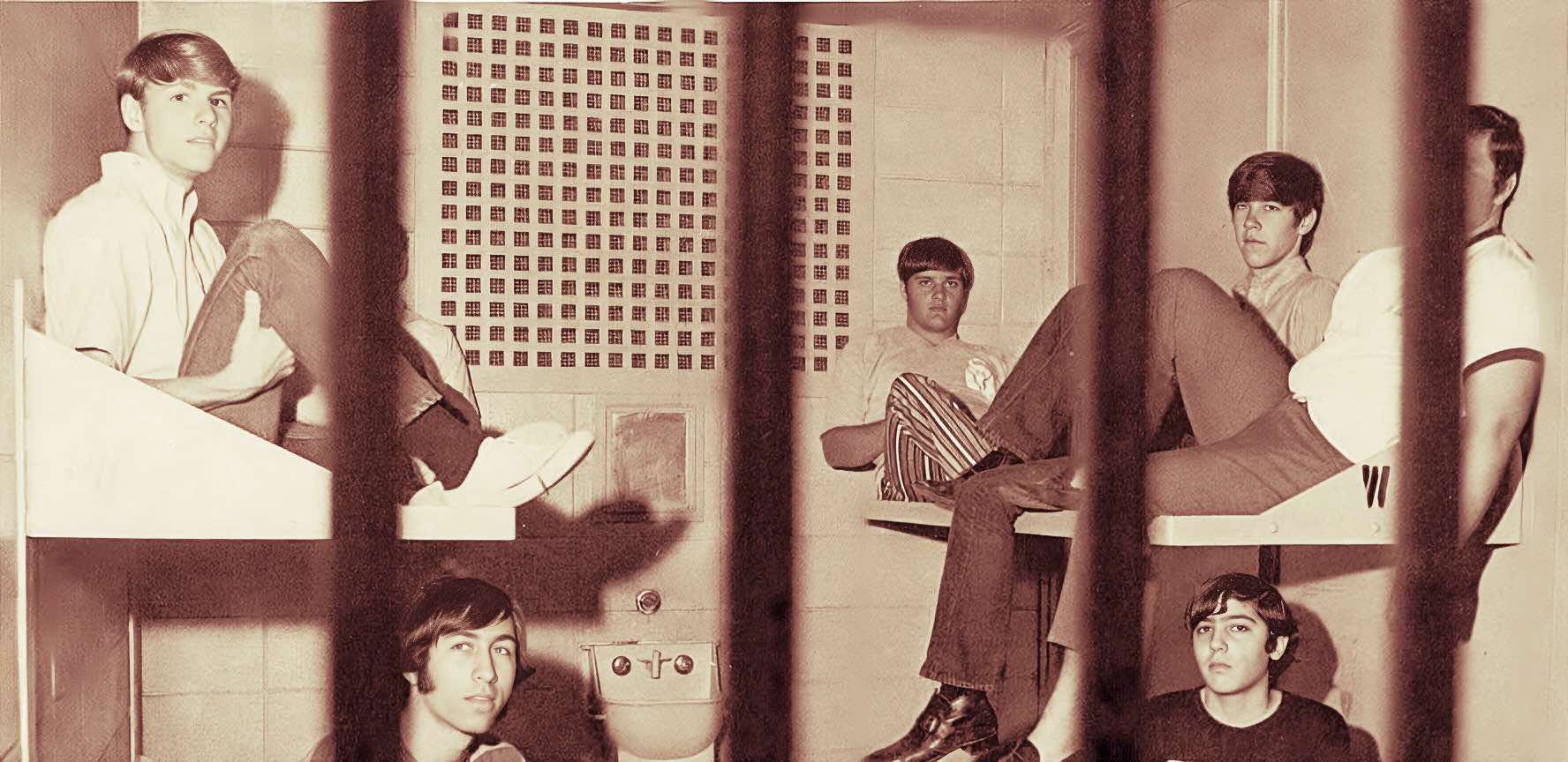 Teenage boys in a vintage prison holding cell, behind bars, with historical context of news, music, and social history archives on Past Daily.