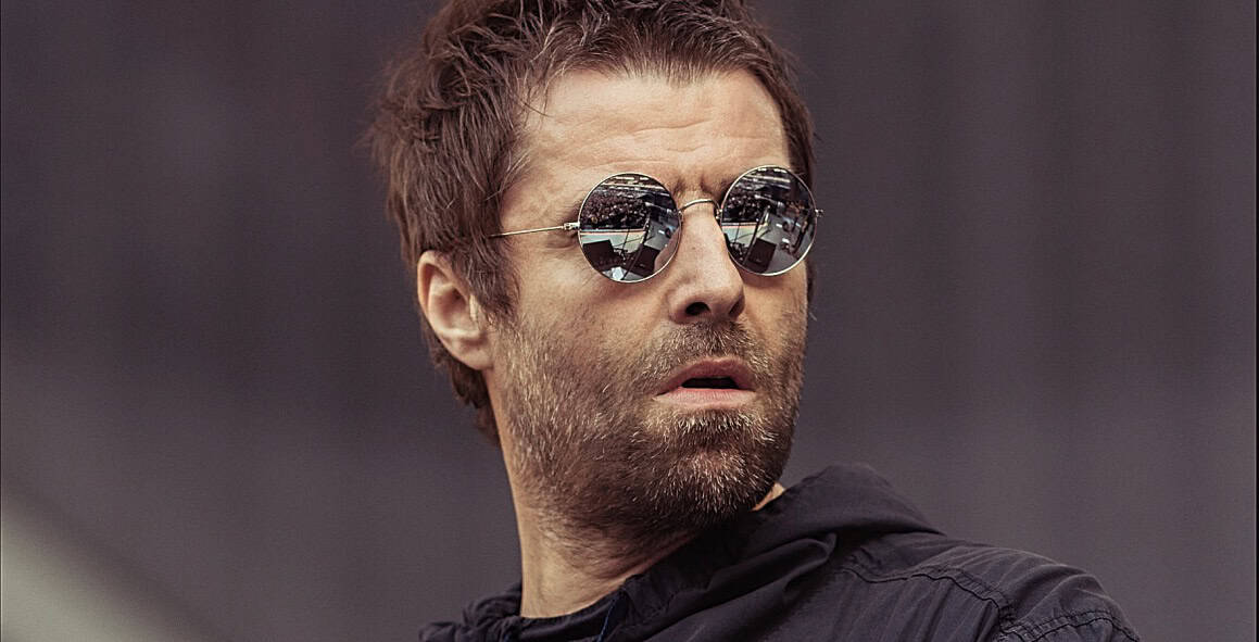 A man with sunglasses and a beard, wearing a black jacket, standing outdoors with a blurred background.