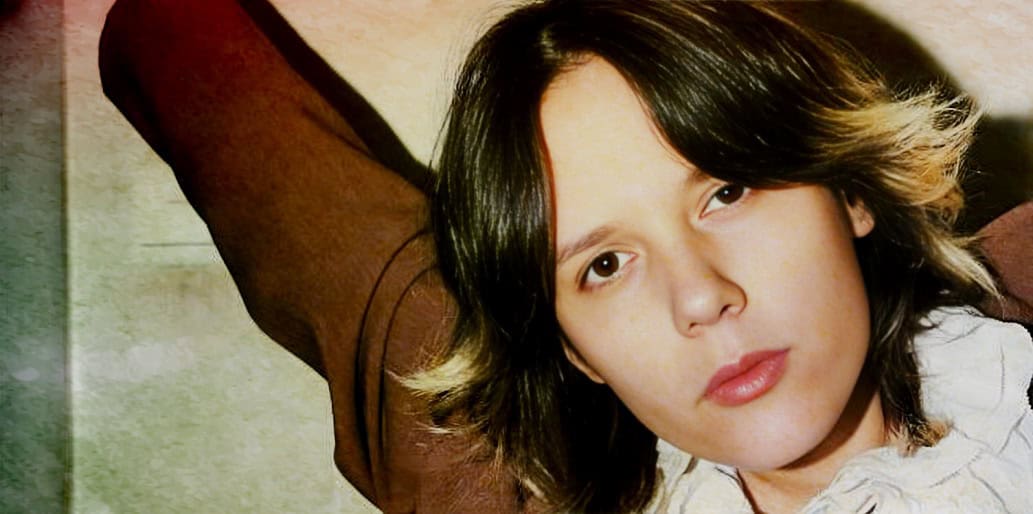 A woman with dark hair and light brown highlights, lying down on a beige surface, looking at the camera, with a relaxed expression behind-the-scenes of a music or radio archive focused on news, history, and music preservation in audio format.