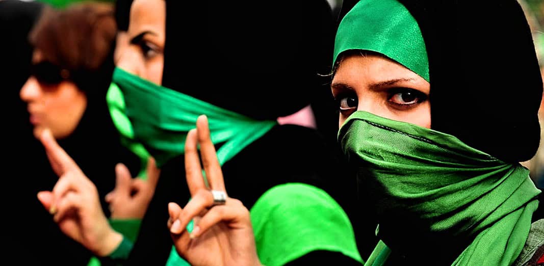 Women wearing green and black hijabs with face coverings, striking a peace sign, in a protest or rally setting, symbolizing activism, women's rights, and social movements.