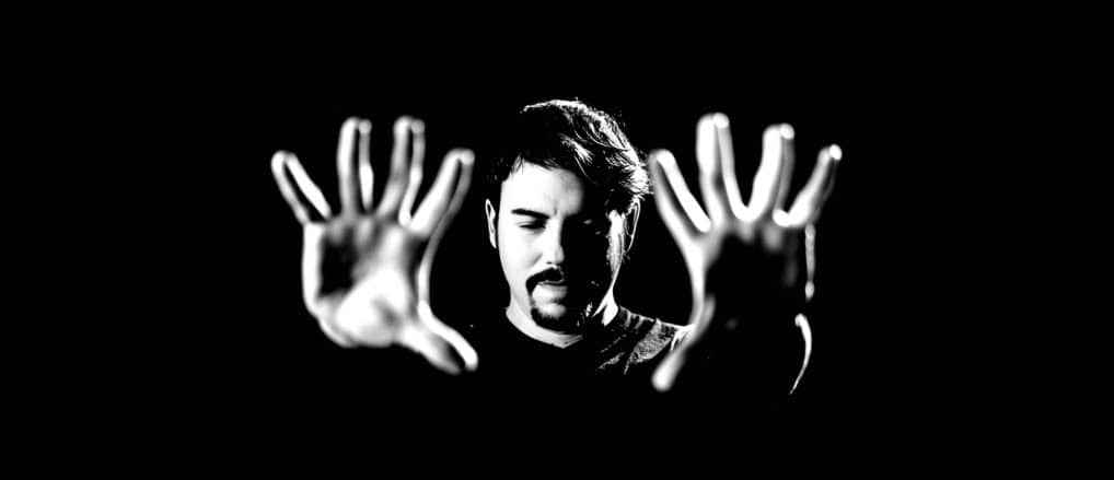 A black and white image of a man with hands extended towards the camera, conveying a dramatic and introspective mood.