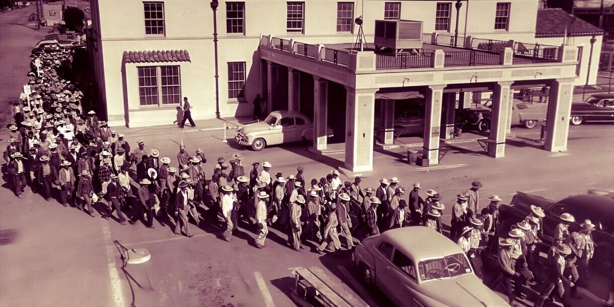 Crowd of people wearing vintage clothing and hats walking on city street, reflective of historical news and music archives from the early 20th century.