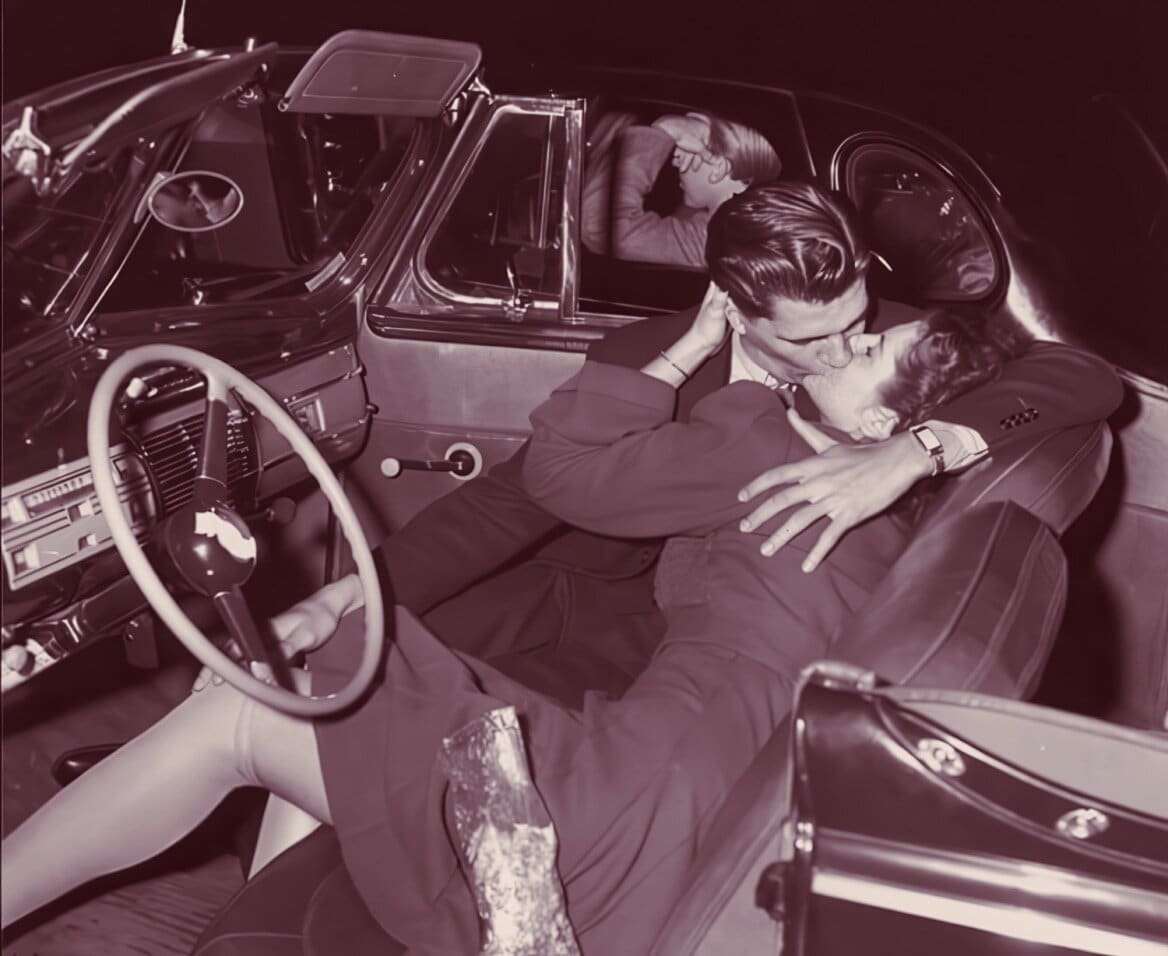 Driving couple kissing inside vintage car, romantic love scene, 1950s style, retro car interior, nostalgic movie scene, classic romance, vintage automobile, historic love story, mid-century vibe, Past Daily archive imagery.