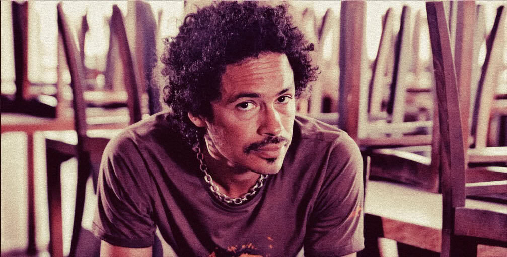 A close-up of a man with curly hair and a mustache, sitting in a room with stacked chairs, representing a podcast or radio host for historical news and music archives.