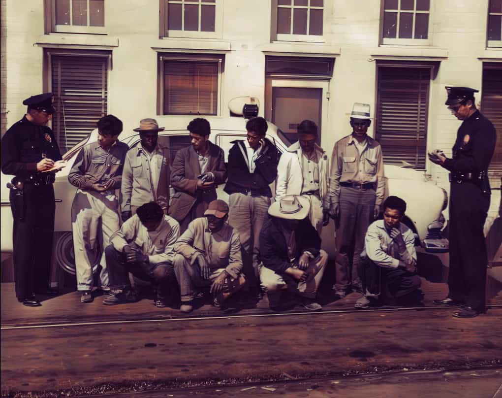 Police officers interviewing a group of men on the street, black and white attire, urban setting, historical scene, vintage photo, reminiscent of past news reports and social history.