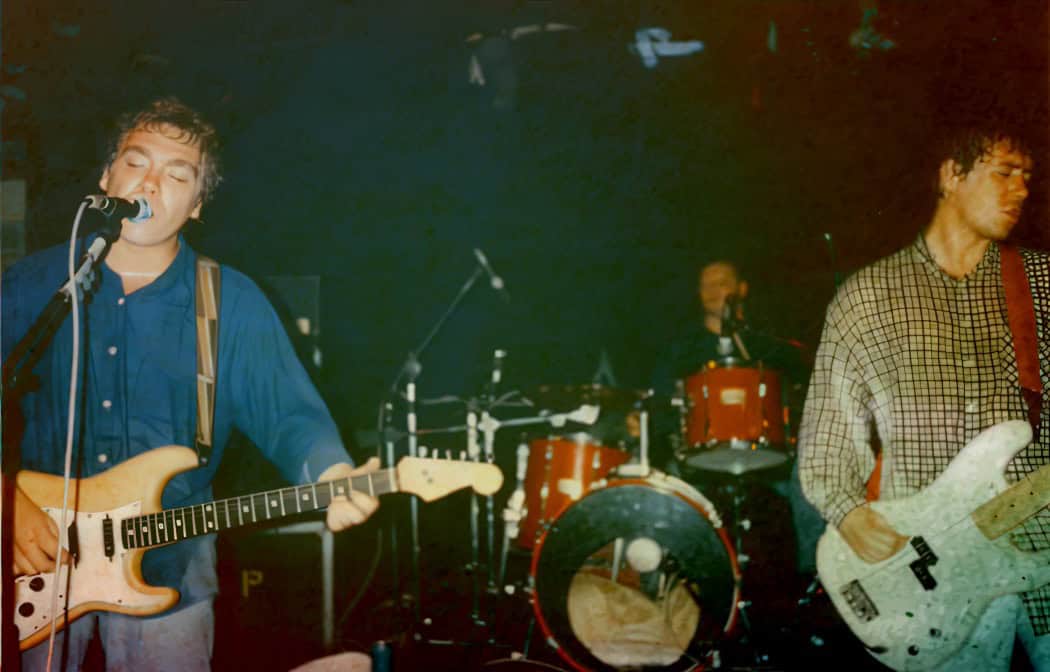 Guitarist singing and playing electric guitar during live concert, band performance with drummer in background, vintage music scene, rock band live show, energetic music event, Past Daily music archive relevance.