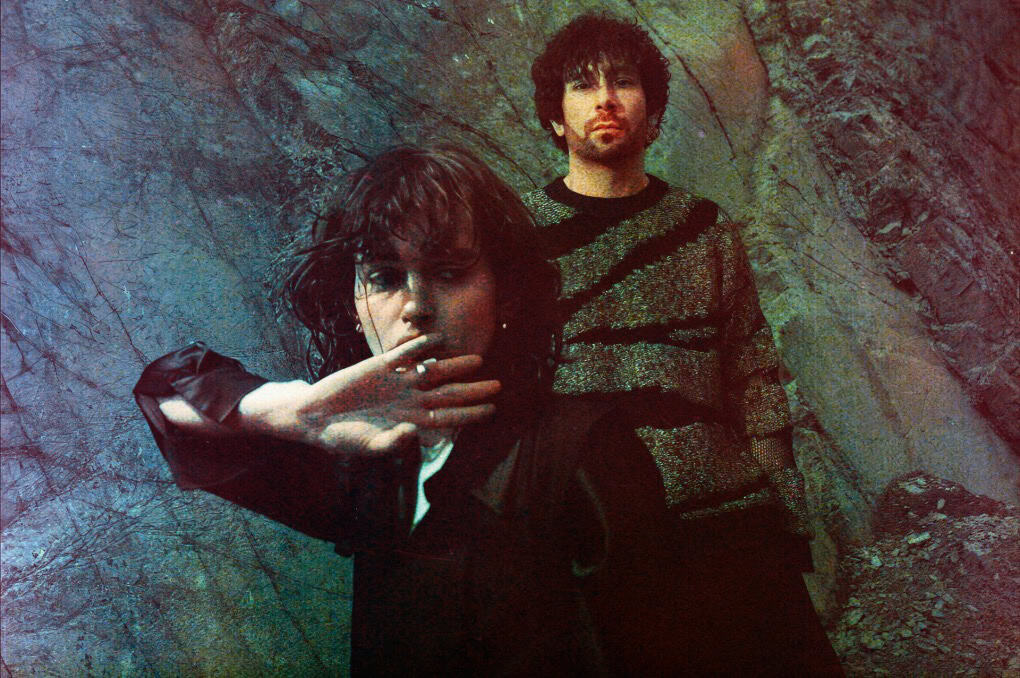 Earth tone patterned clothing, artistic portrait of a man and woman, moody outdoor setting, web archive for news, history, and music history, Past Daily sound archive, vintage style, alternative fashion, stylish music duo in natural environment.