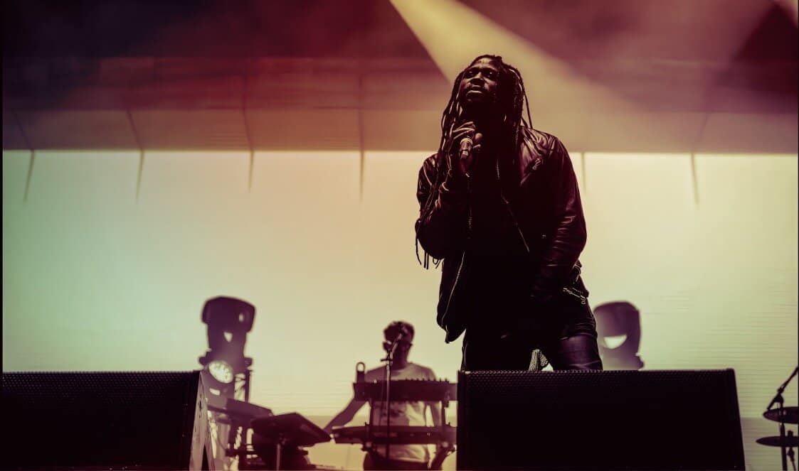 Vocalist with dreadlocks singing on stage during live music performance, illuminated by stage lights, with band members and musical equipment in background, capturing the essence of live concert music.