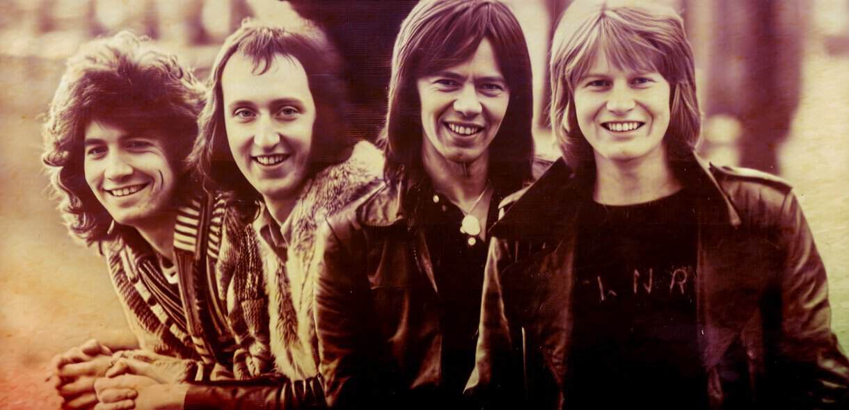A vintage photo of four young men with 1970s hairstyles and leather jackets, representing a classic rock band from the era.