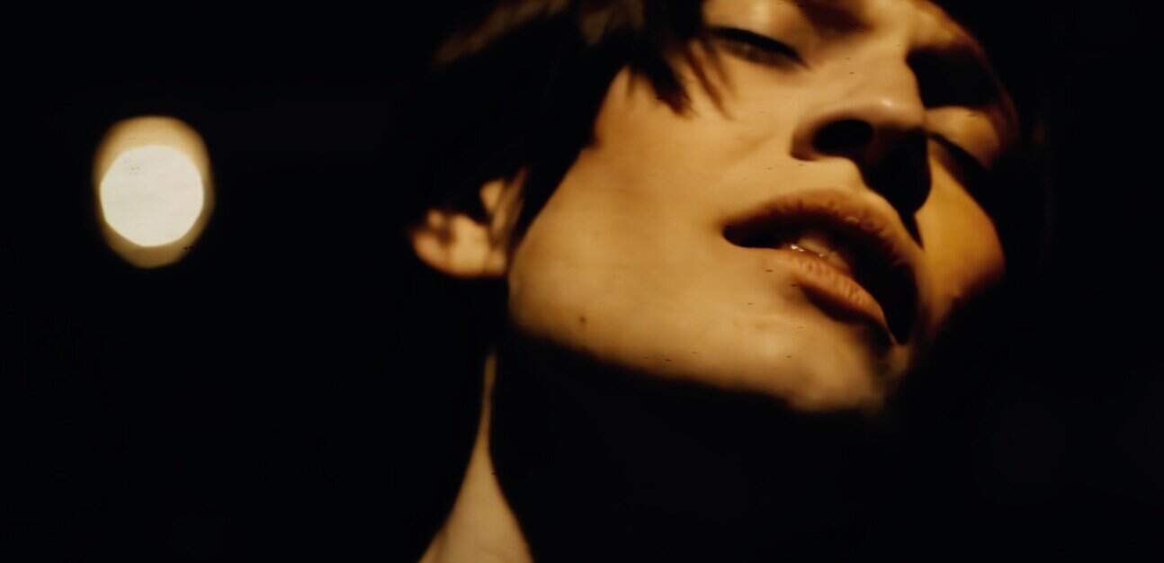 A woman with short hair, closed eyes, and face paint in a dark, moody setting, illuminated by soft light, capturing an artistic and expressive moment.