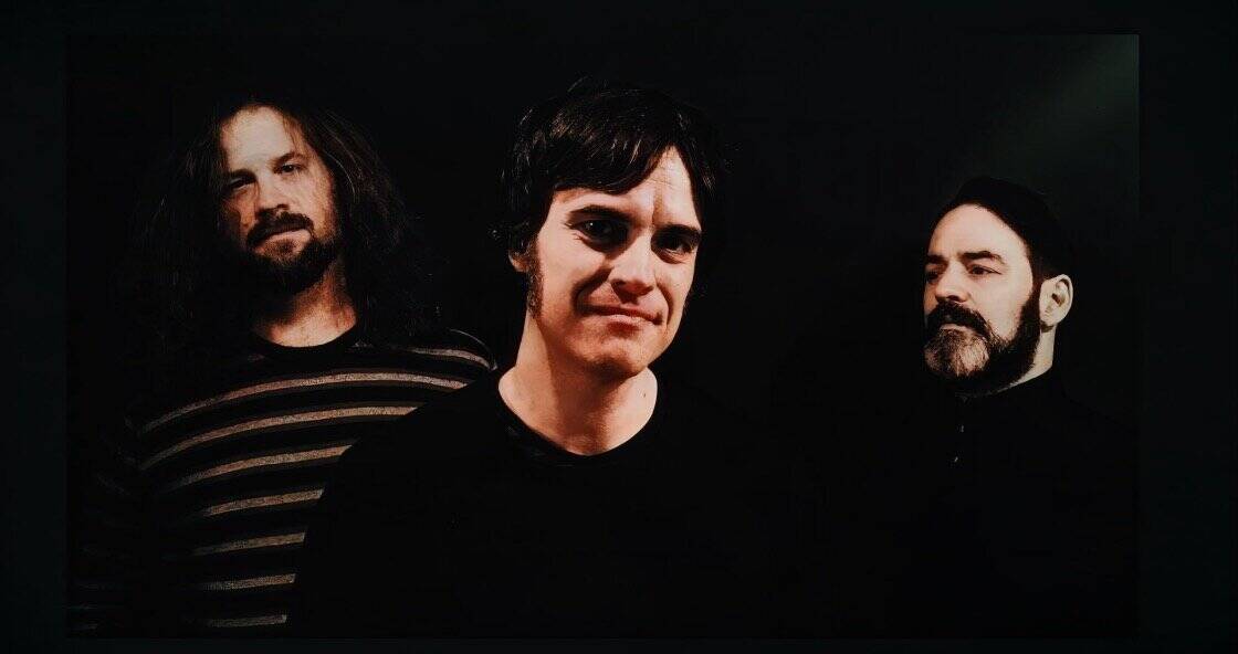 Vintage rock band members with dark hair and beards, styled in black clothing, against a black background, representing classic music history and sound archives.
