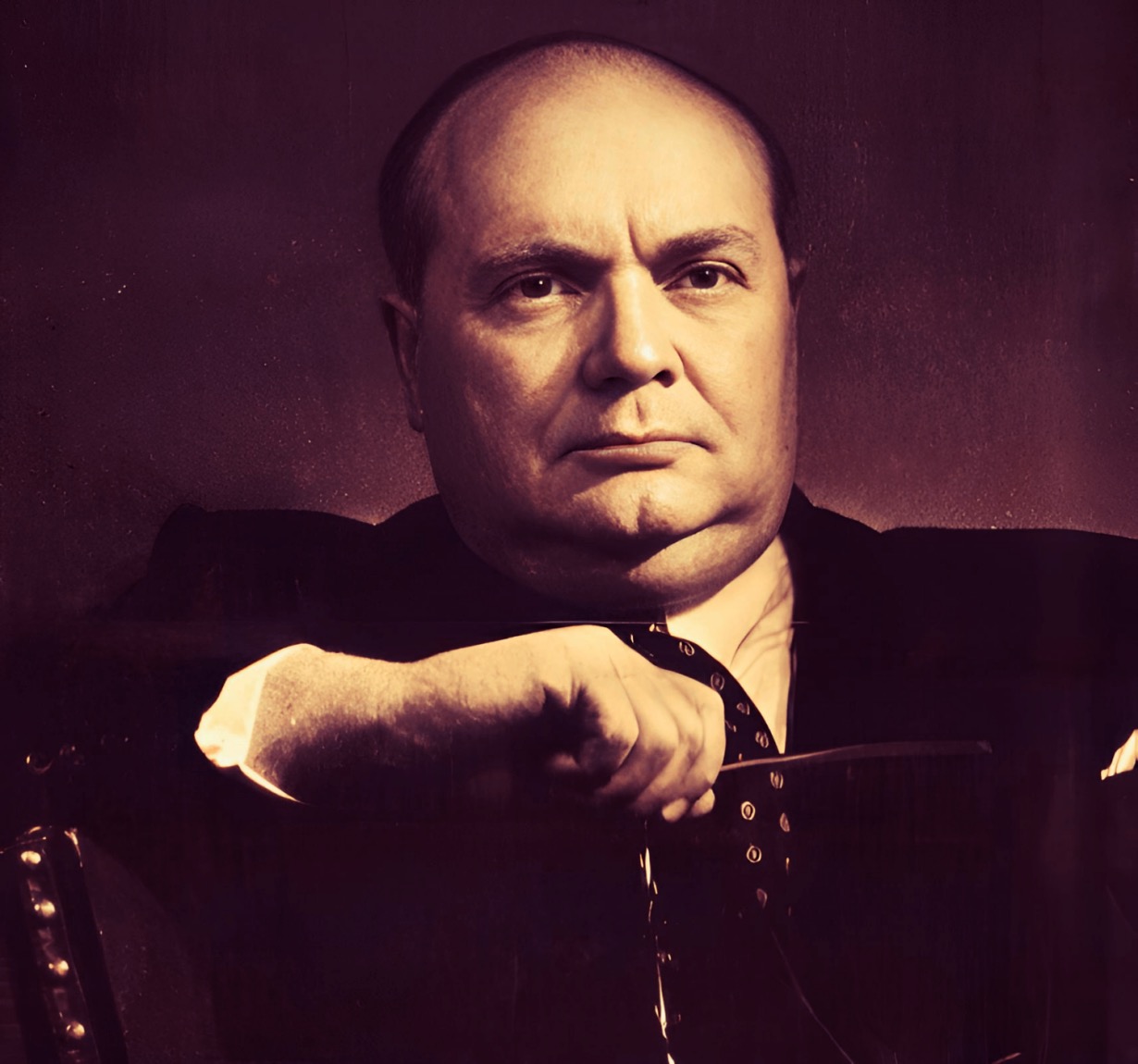 A man with a serious expression, wearing a suit and tie, sitting with his hand resting on a chair. The background is dark, emphasizing his facial features and poised demeanor. This image is related to news, history, and music archives.