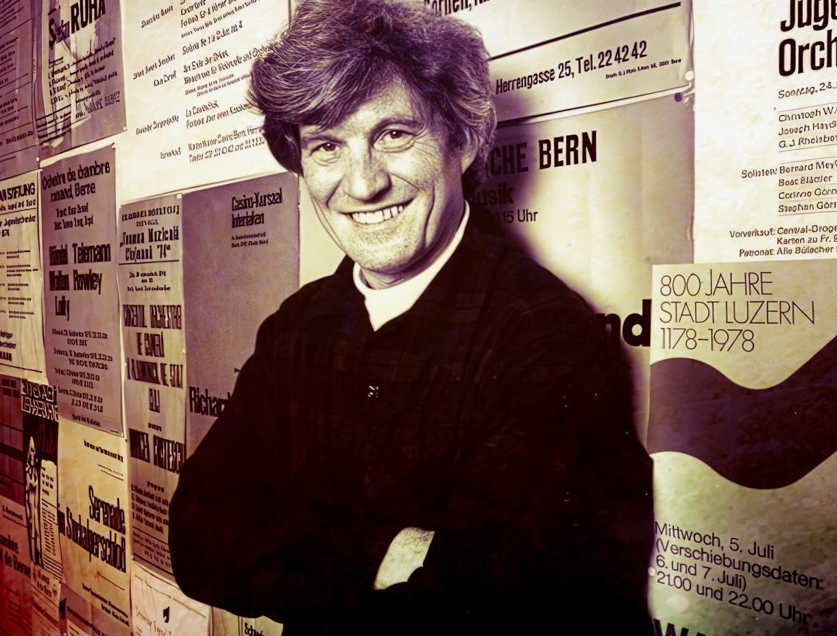 A smiling man with light-colored hair standing in front of a wall covered with various event posters and announcements.