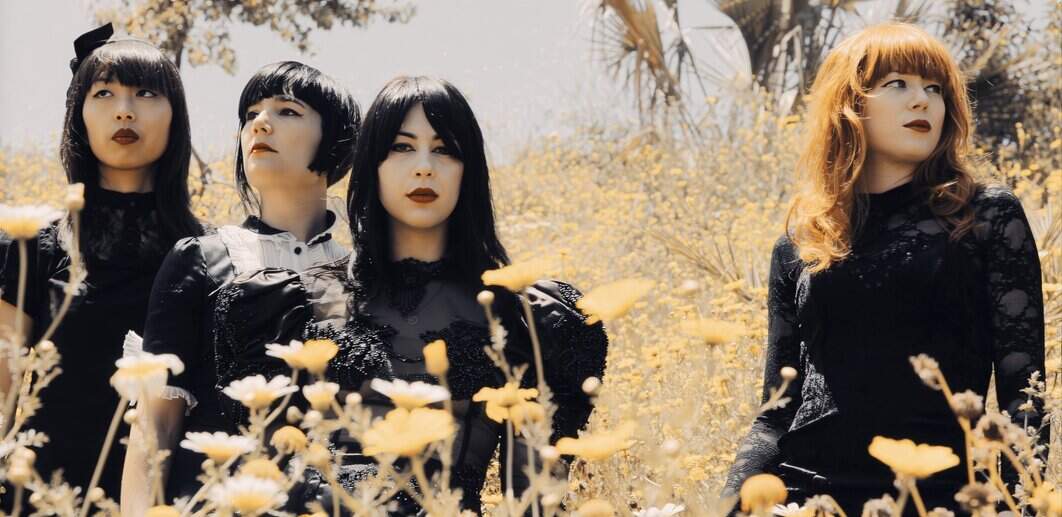 Elegant women dressed in black with contrasting hair colors standing amidst golden wildflowers in a natural outdoor setting.