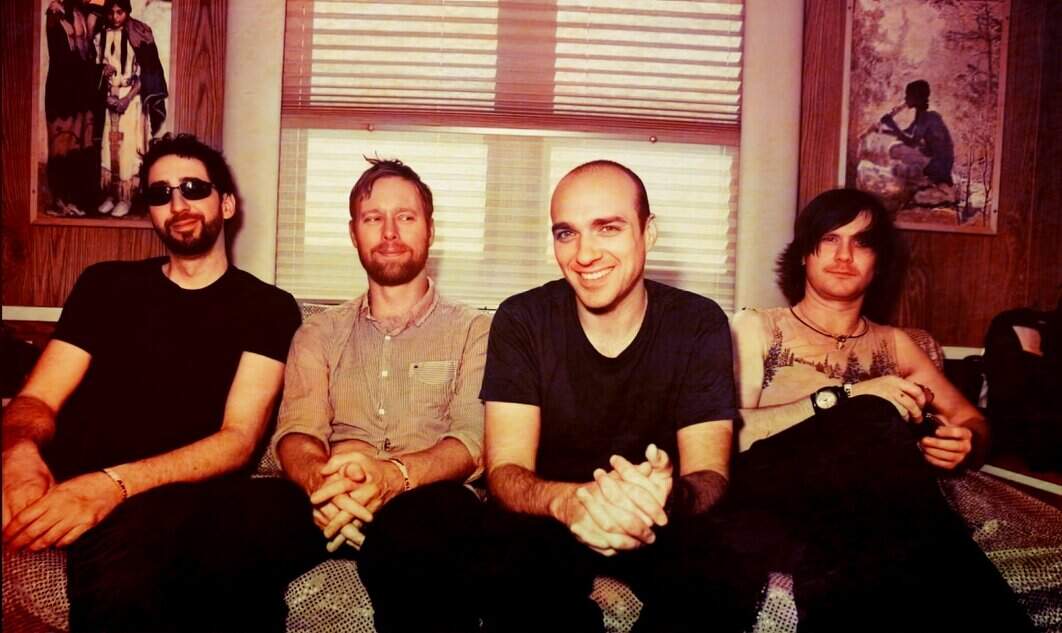 Dream pop band sitting on a couch in a cozy, vintage-styled room with wooden walls and framed artwork, smiling and relaxed, creating a warm, inviting vibe for music and nostalgia enthusiasts.