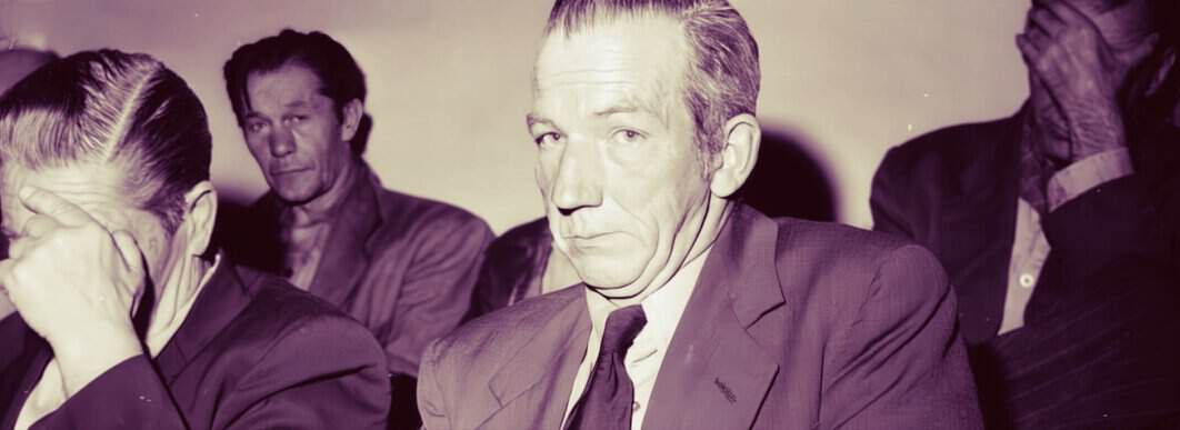 Somber black-and-white image of men in suits, including a serious-looking older man, possibly in a courtroom or formal hearing, reflecting historical news and legal themes.