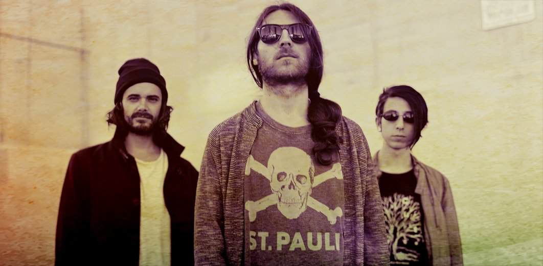 Dark-haired band members wearing sunglasses and casual clothing, standing outdoors against a beige wall, representing a music group or rock band scene.