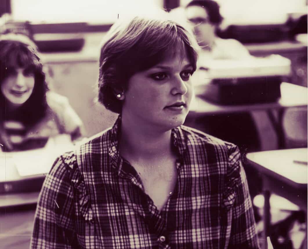 Young woman with short hair in a classroom, focused and attentive during a lecture, with other students visible in the background.