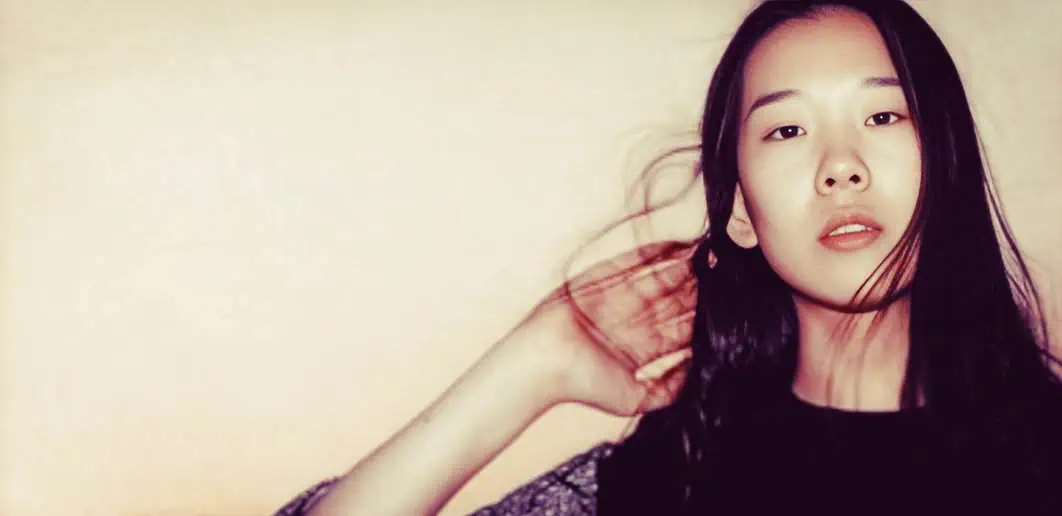 A young Asian woman with long black hair posing against a neutral background, capturing a contemplative and stylish expression.