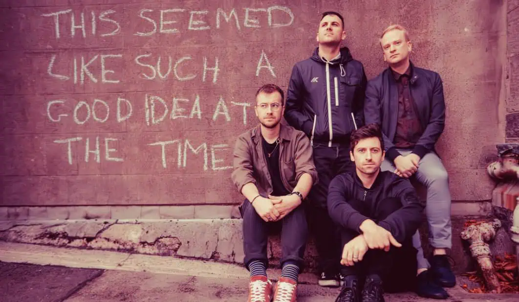 A band of four young men posing against a graffiti wall with message writing, capturing a modern, artistic vibe associated with music and cultural expression.