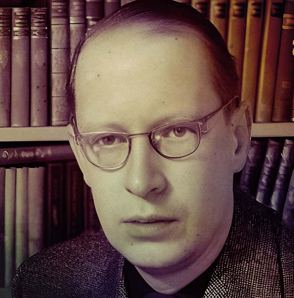 Vintage black and white portrait of a man with glasses, set against a background of bookshelves filled with old books, evoking a sense of history and scholarly research.