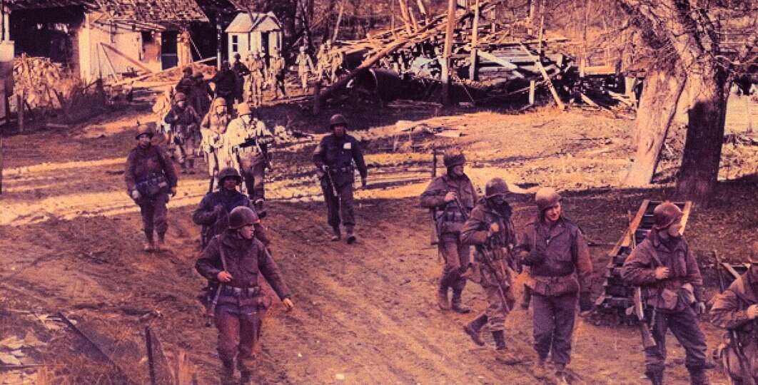 Armed soldiers walking through a war-torn neighborhood with destroyed houses and debris in the background.