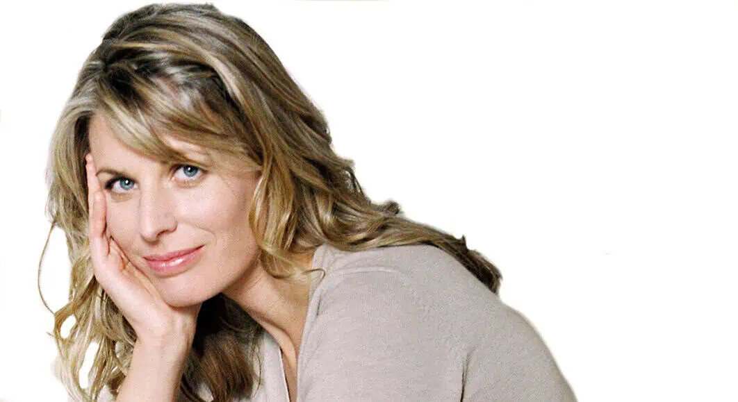 Close-up portrait of a smiling woman with blonde hair, resting her face on her hand, set against a plain white background.