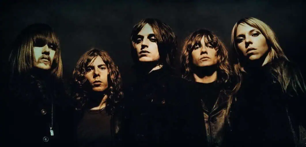 Rock band members in dark leather jackets, posing seriously against a black background, representing classic rock or alternative music scene, with intense expressions and layered hairstyles.