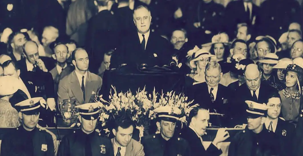 A historical black-and-white photograph of Franklin D. Roosevelt delivering a speech at a formal event, surrounded by an attentive audience of men and women, many wearing hats, during the era of the New Deal.