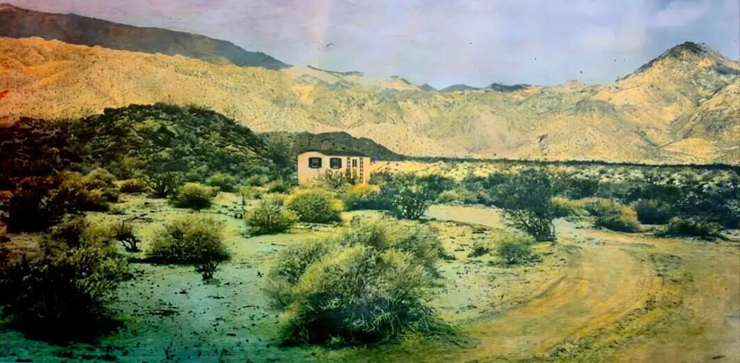 Desert landscape with sparse bushes and a small modern building at the base of distant mountains in a remote area.