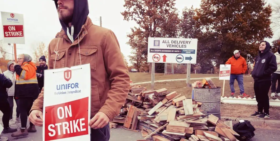 Unifor workers on strike protesting labor dispute with signs and broken wood debris at picket line, outdoor setting with others in background near signage and trees, labor rally image.