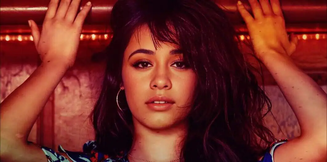 Women with long dark hair posing indoors, head tilted, hands raised near her head, wearing hoop earrings, vibrant lighting and textured background.
