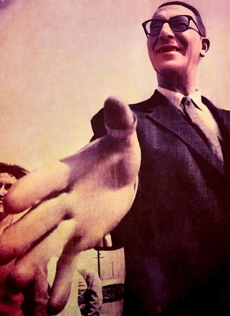 A man in a suit and glasses smiling and pointing toward the camera, with a pig held close to his chest, creating a humorous and quirky scene.