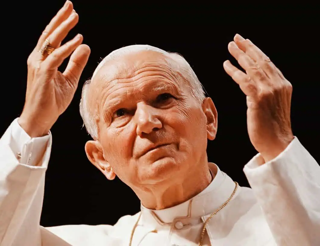 Elderly man with white hair making a gesture with both hands, wearing a white robe and gold necklace.