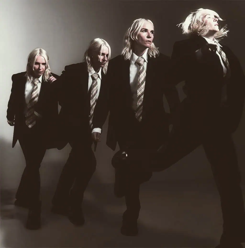 Blonde women in black suits with plaid ties, performing a choreographed dance in a minimalistic studio setting.
