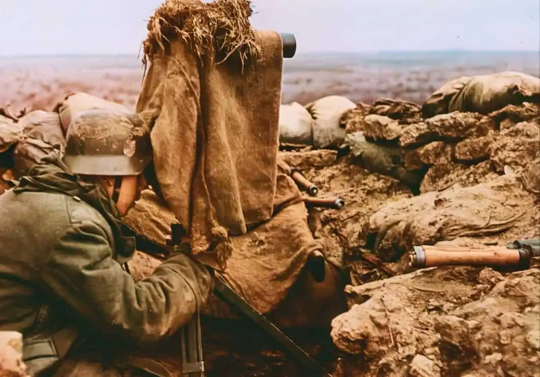U.S. soldier in trench during combat, wearing helmet and camouflage, aiming rifle from a fortified position in a war zone, with a rugged landscape in the background.