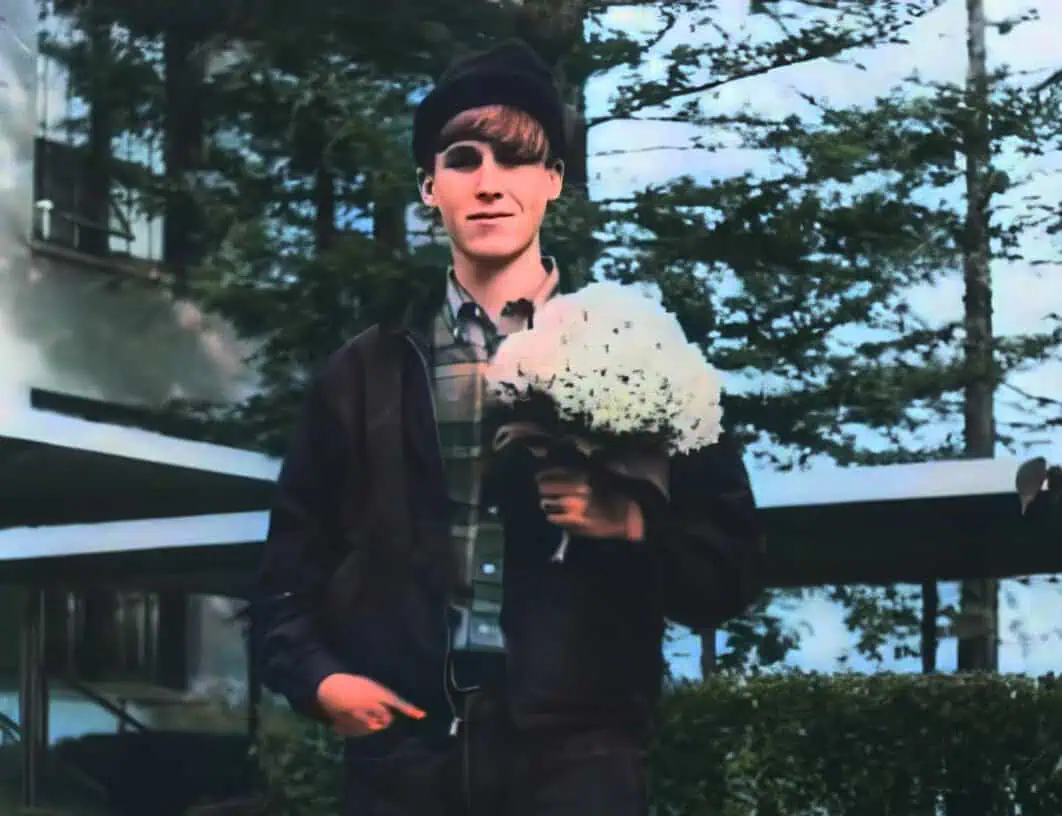 Young man holding a bouquet of white flowers outdoors in front of trees and a modern building, wearing a black jacket, plaid shirt, and black hat.